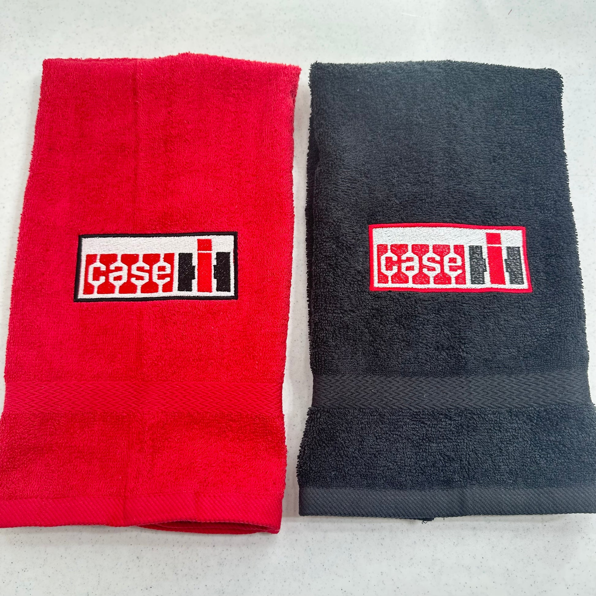 Two folded towels side by side on a light gray surface: a red towel on the left and a dark charcoal gray towel on the right, each with a rectangular embroidered Case IH logo patch on the front.