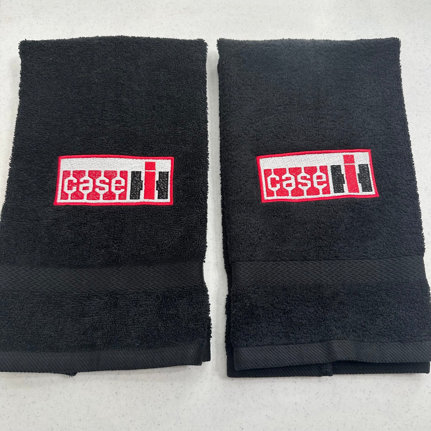 Two black towels laid flat side by side on a light gray surface, each with a rectangular red, white, and black embroidered patch featuring the word 'case' in stylized lettering.