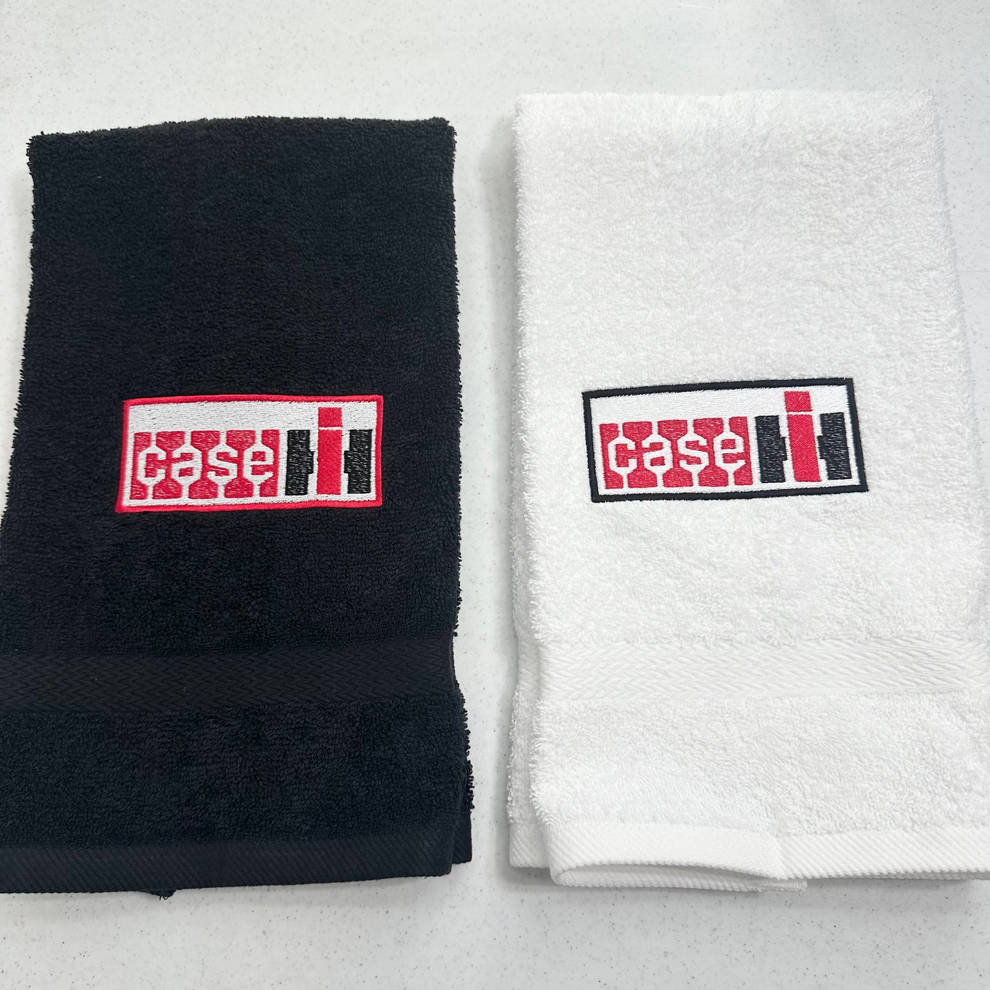 Two folded towels side by side on a light gray surface: a black towel on the left and a white towel on the right, each with a rectangular embroidered Case IH logo patch on the front.