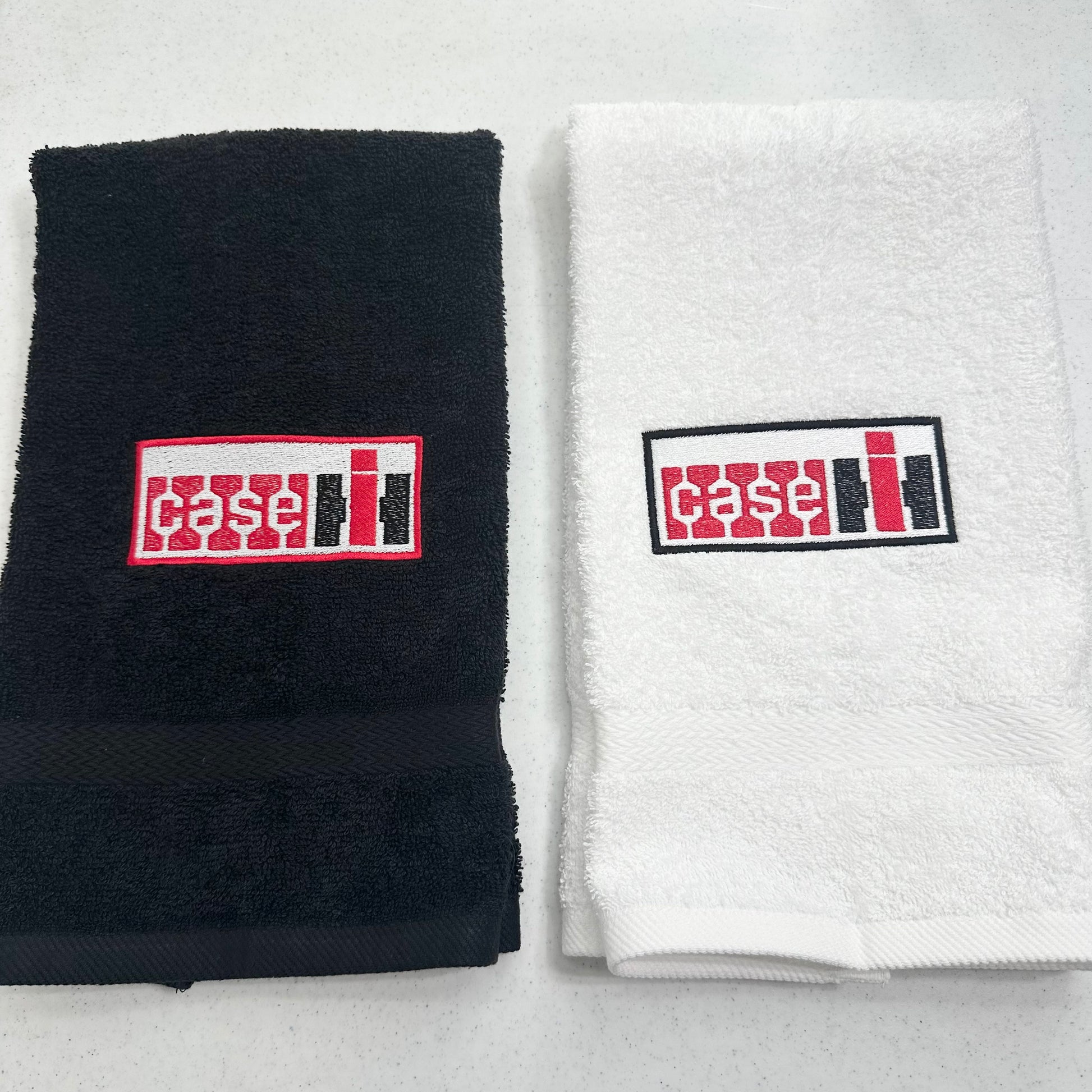 Two folded towels side by side on a light gray surface: a black towel on the left and a white towel on the right, each with a rectangular embroidered Case IH logo patch on the front.