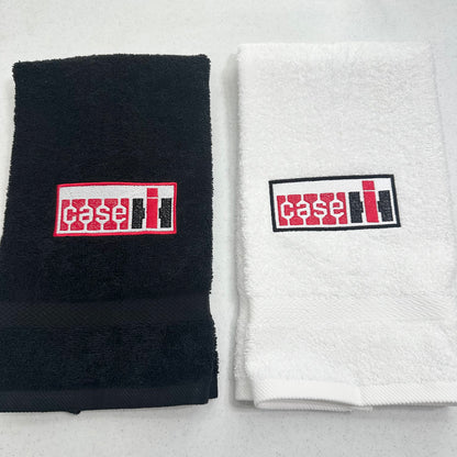 Two folded towels side by side on a light gray surface: a black towel on the left and a white towel on the right, each with a rectangular embroidered Case IH logo patch on the front.