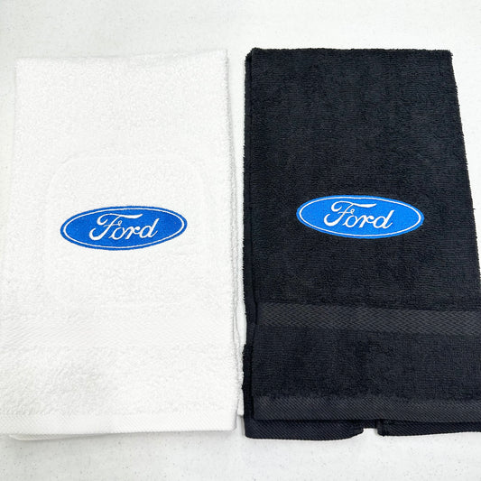 Two folded towels side by side on a white background, one white and one black, each featuring an embroidered blue oval Ford logo on the front.