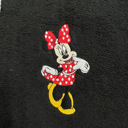 A close-up of a black textured fabric, likely a towel, featuring an embroidered Minnie Mouse character in her red and white polka-dot dress with yellow shoes and a red polka-dot bow.