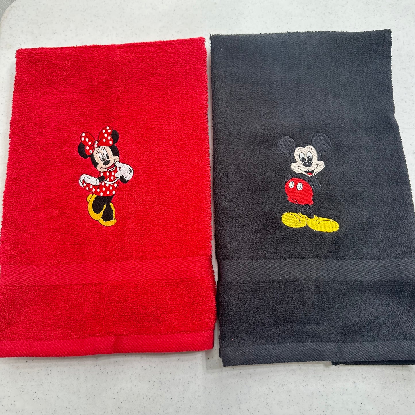 Two folded towels side by side on a light gray surface: a red towel with an embroidered Minnie Mouse motif and a dark charcoal towel with an embroidered Mickey Mouse motif.