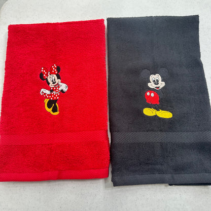 Two folded towels side by side on a light gray surface: a red towel with an embroidered Minnie Mouse motif and a dark charcoal towel with an embroidered Mickey Mouse motif.