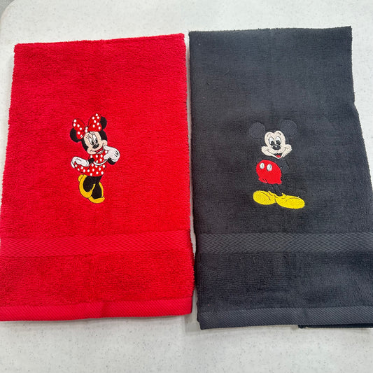 Two folded towels side by side on a light gray surface: a red towel with an embroidered Minnie Mouse motif and a dark charcoal towel with an embroidered Mickey Mouse motif.