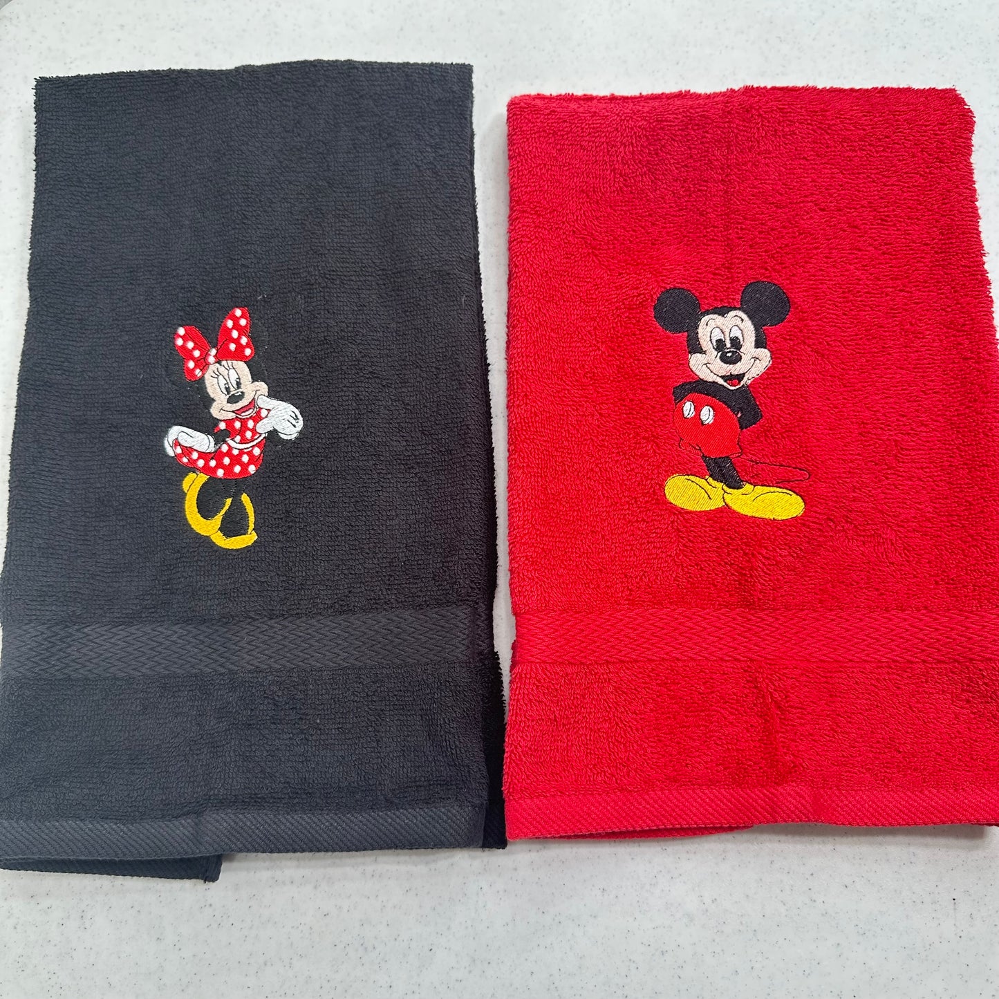 Two folded cotton towels side by side on a light gray surface: one black towel with an embroidered Minnie Mouse motif, and one red towel with an embroidered Mickey Mouse motif.