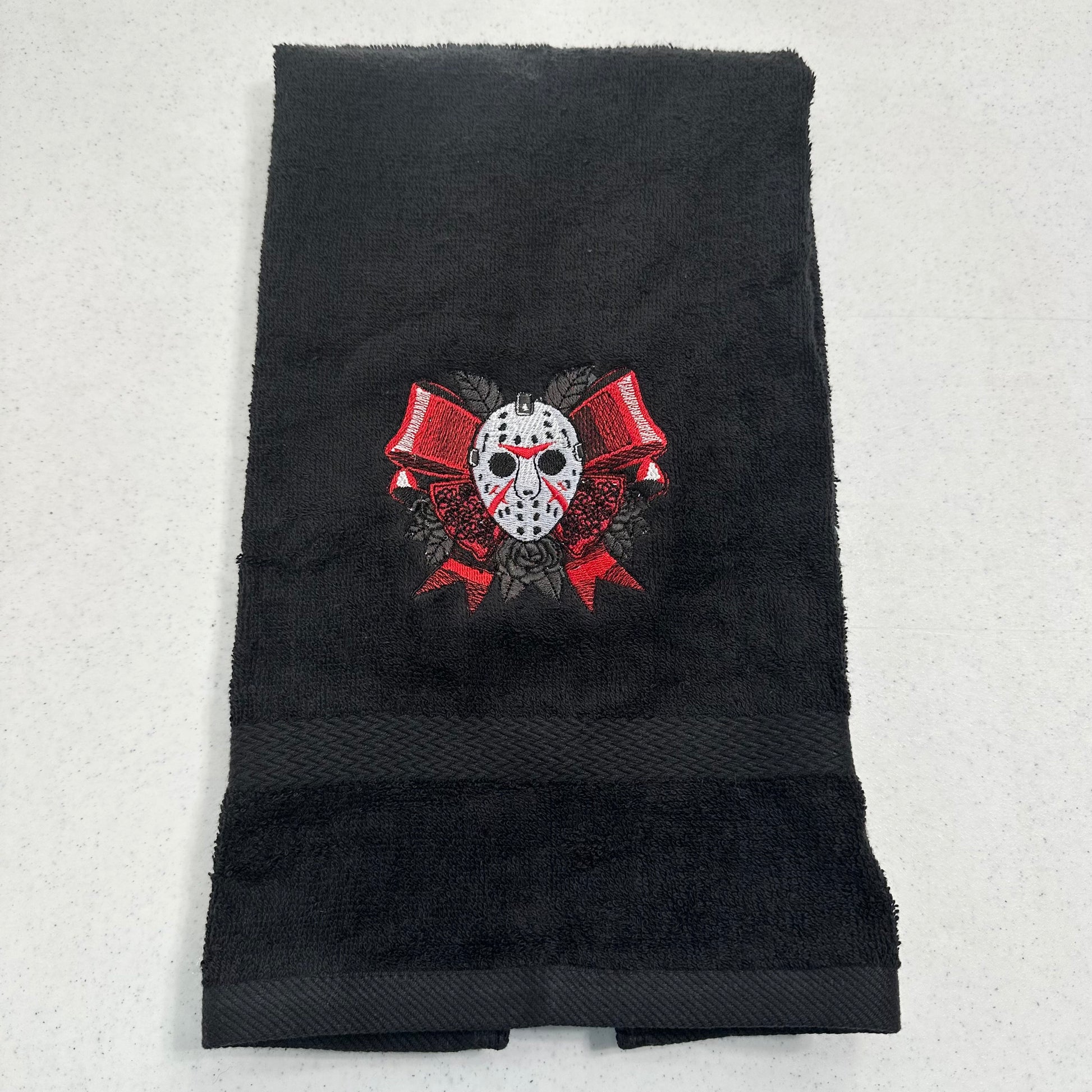 A black towel featuring an embroidered graphic of jason hockey mask with red bows and roses, laid flat on a light gray background.