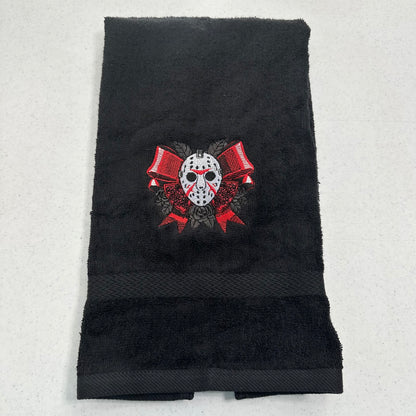 A black towel featuring an embroidered graphic of jason hockey mask with red bows and roses, laid flat on a light gray background.
