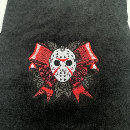 A close-up of a black textured fabric, likely a towel or blanket, featuring a central embroidered graphic of a white hockey mask with red markings, flanked by red and black roses and ribbons.