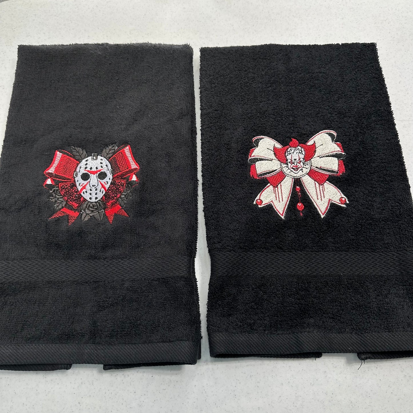 Two folded black towels on a light gray background, each with a large embroidered horror-themed bow motif. The left towel shows a hockey mask under red ribbons, and the right towel shows a pennywise clown face under a white-and-red bow.
