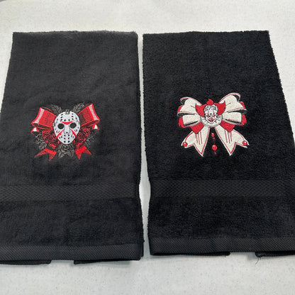 Two folded black towels on a light gray background, each with a large embroidered horror-themed bow motif. The left towel shows a hockey mask under red ribbons, and the right towel shows a pennywise clown face under a white-and-red bow.