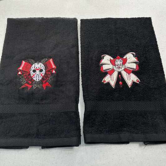 Two folded black towels on a light gray background, each with a large embroidered horror-themed bow motif. The left towel shows a hockey mask under red ribbons, and the right towel shows a pennywise clown face under a white-and-red bow.
