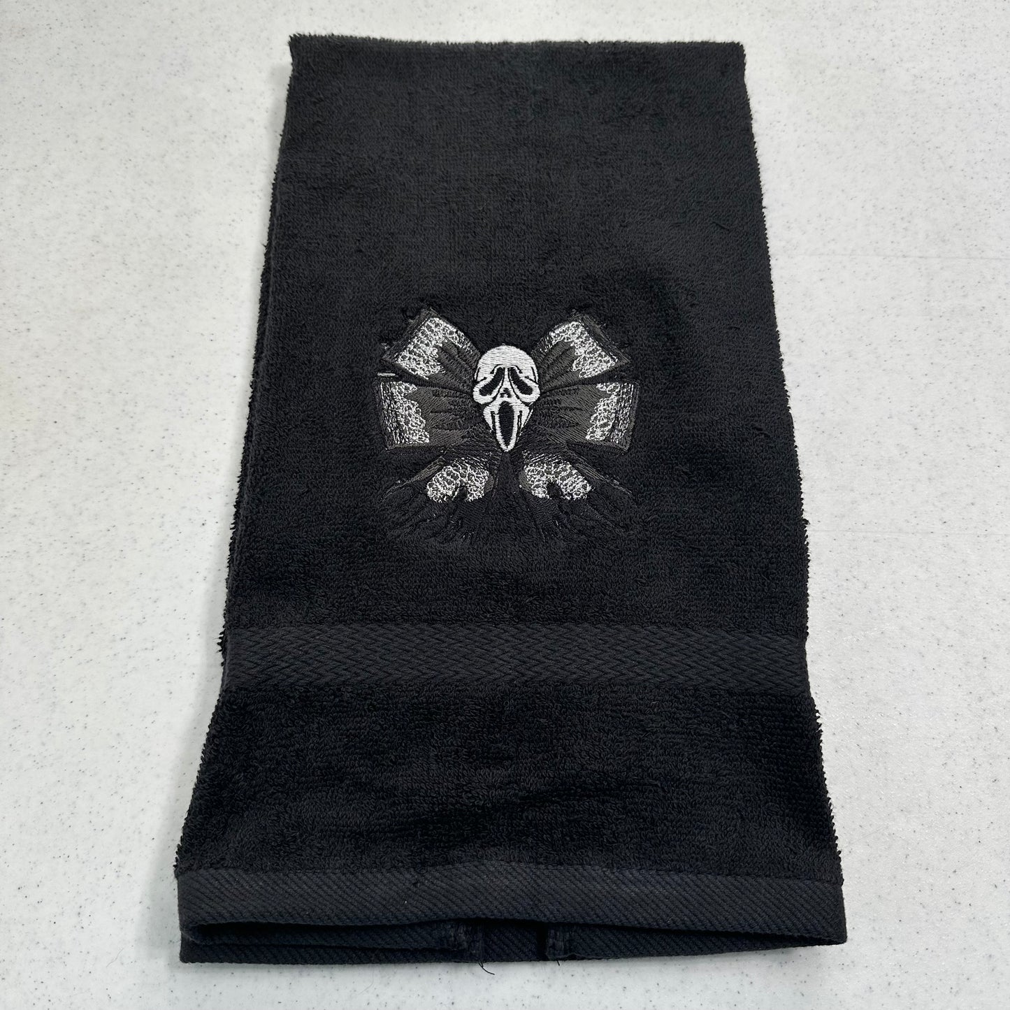 A black terry cloth towel featuring an embroidered Ghostface mask and crossed bones motif in the center, laid flat on a light gray textured surface.