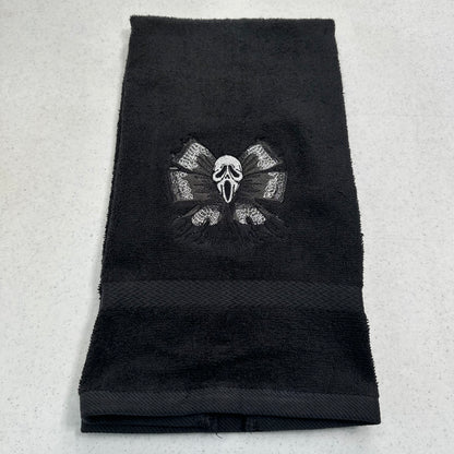 A black terry cloth towel featuring an embroidered Ghostface mask and crossed bones motif in the center, laid flat on a light gray textured surface.