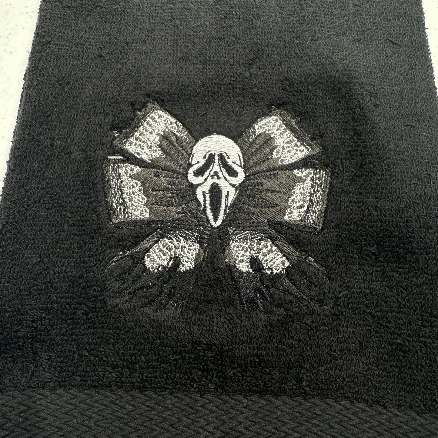 A close-up of a black textured towel featuring an embroidered design of the Ghostface mask from the Scream franchise, flanked by two distressed gray and black wings.