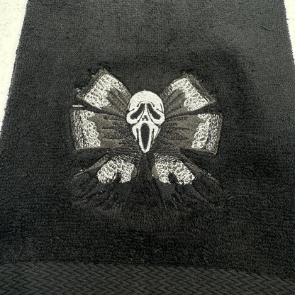 A close-up of a black textured towel featuring an embroidered design of the Ghostface mask from the Scream franchise, flanked by two distressed gray and black wings.