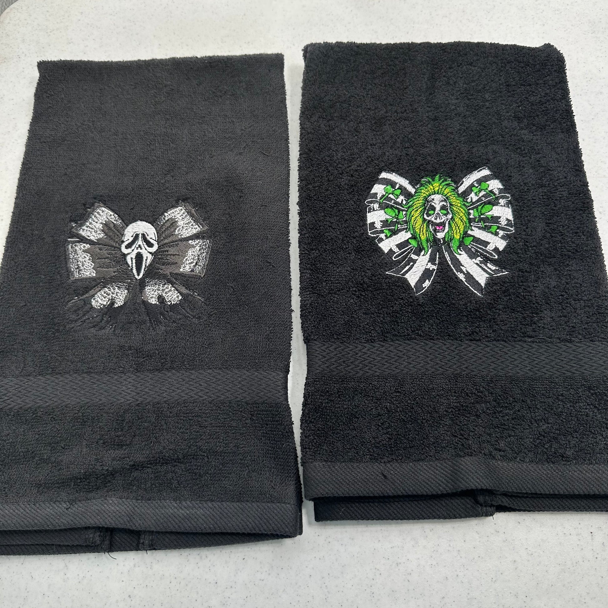 Two black towels laid flat on a light gray surface, each featuring a stitched horror-themed skull and ribbon motif. The towel on the left has a dark gray ghostface design, and the towel on the right has a green-and-white beetlejuice skull design.