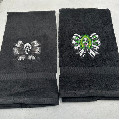 Two black towels laid flat on a light gray surface, each featuring a stitched horror-themed skull and ribbon motif. The towel on the left has a dark gray ghostface design, and the towel on the right has a green-and-white beetlejuice skull design.