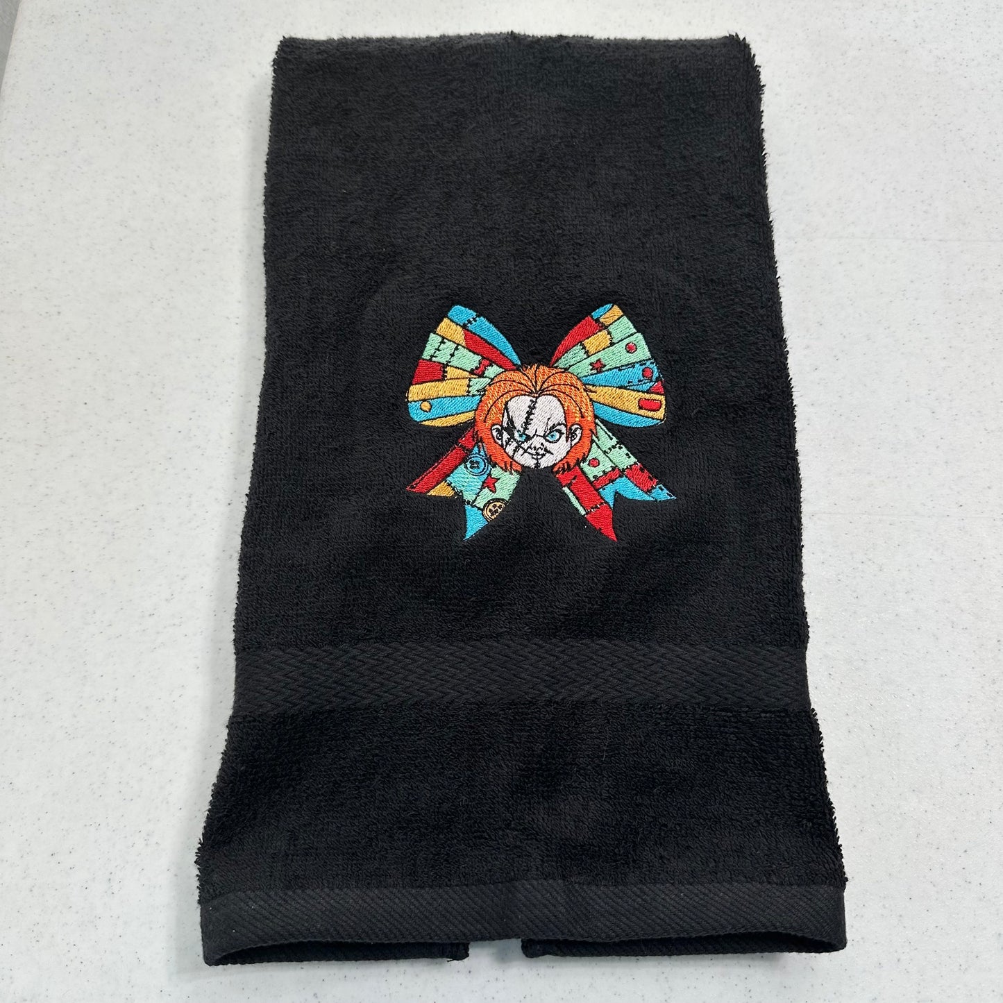 A black cotton towel featuring an embroidered patch of a stylized chucky character with a colorful mosaic bow, displayed on a light gray textured surface.