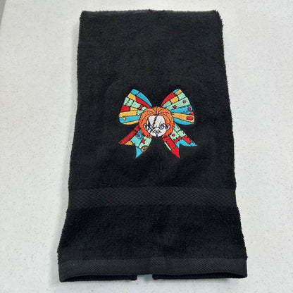 A black cotton towel featuring an embroidered patch of a stylized chucky character with a colorful mosaic bow, displayed on a light gray textured surface.