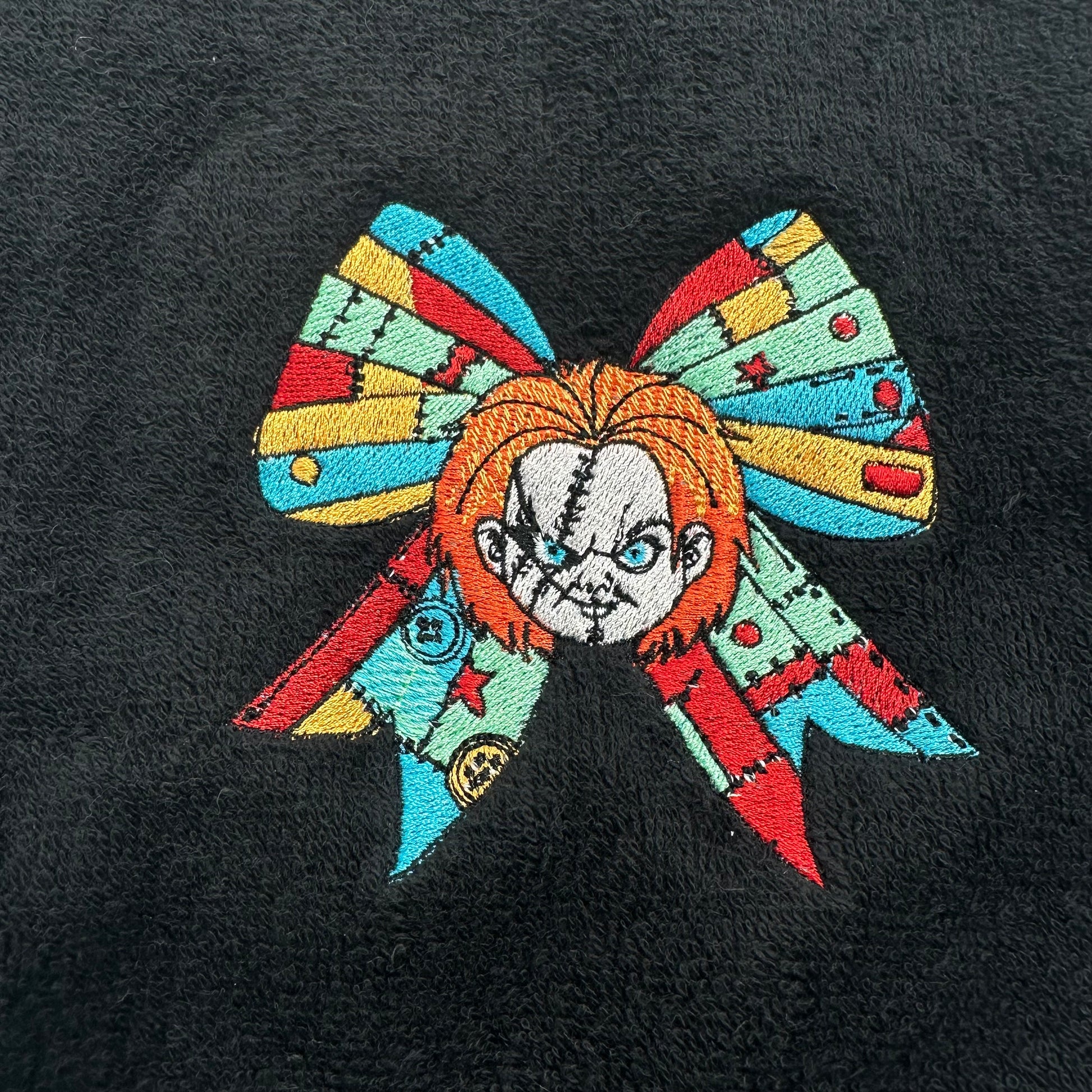 A close-up of a colorful embroidered bow graphic on a black textured fabric background. The bow features a patchwork design in red, yellow, light blue, and mint green, with a stylized stitched face in the center.