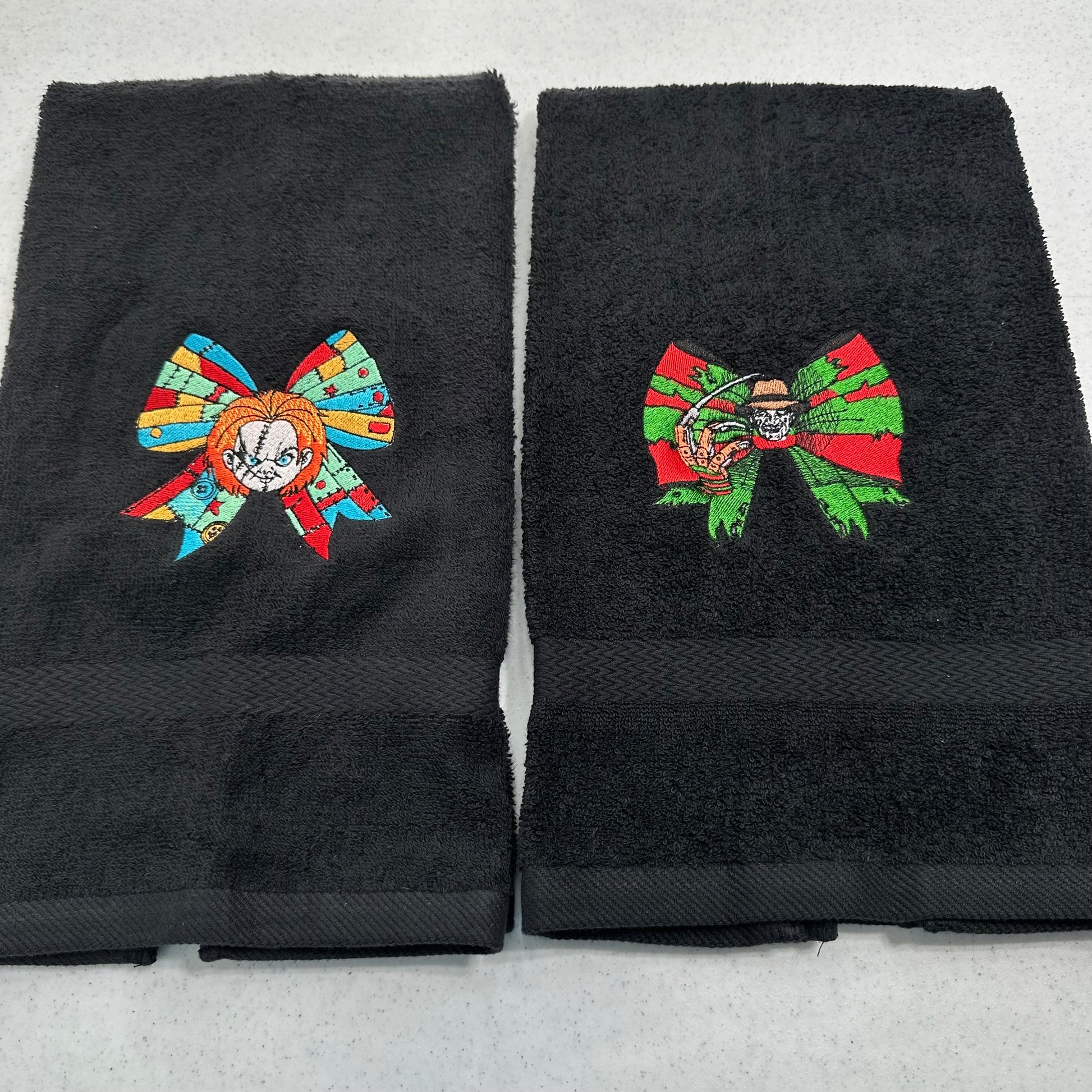 Two black towels side by side, each featuring a colorful embroidered bow design with a chucky character inside. The left bow contains orange, blue, and red details, and the right bow contains green, red, and orange details.