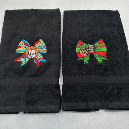 Two black towels side by side, each featuring a colorful embroidered bow design with a chucky character inside. The left bow contains orange, blue, and red details, and the right bow contains green, red, and orange details.