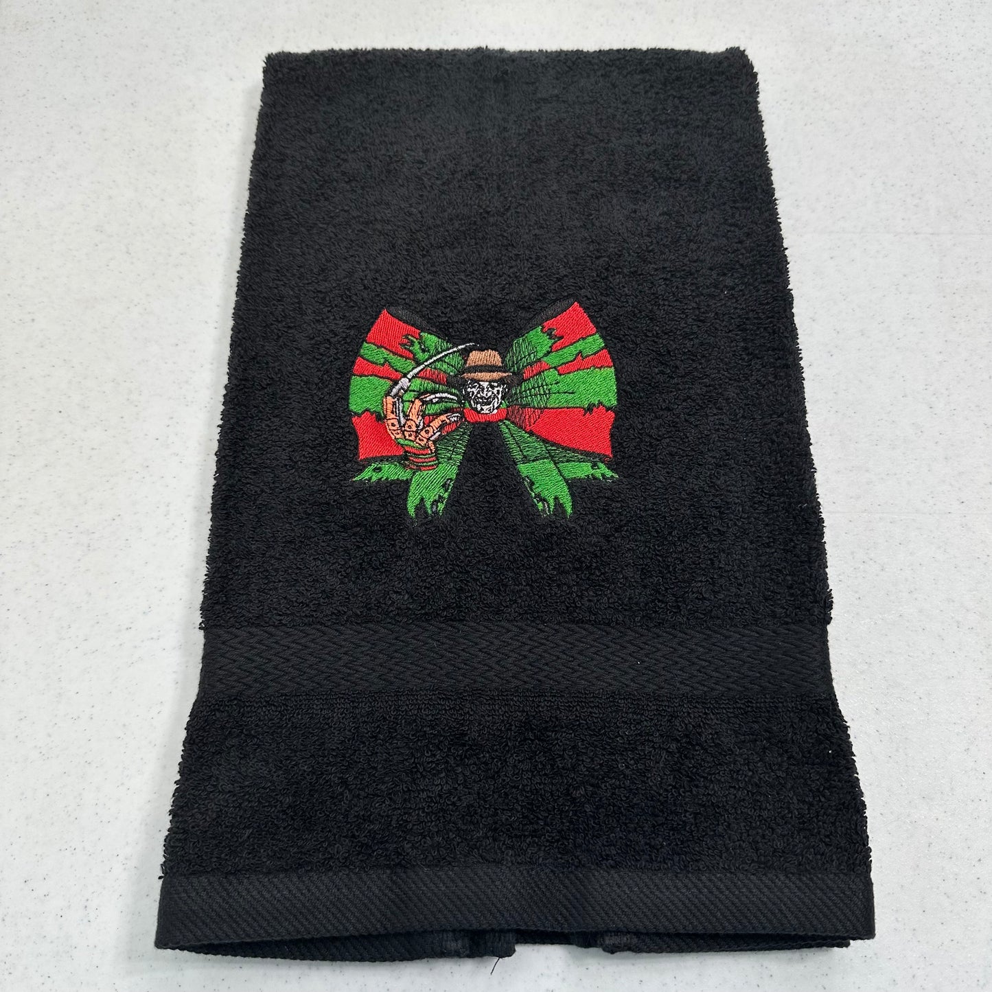 A black terry cloth hand towel featuring an embroidered design of a green and red bow with freddy krueger in the center, laid flat on a light gray surface.