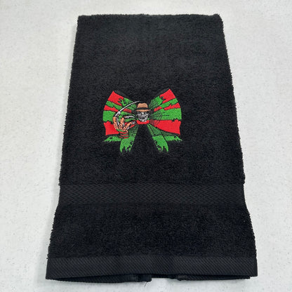 A black terry cloth hand towel featuring an embroidered design of a green and red bow with freddy krueger in the center, laid flat on a light gray surface.