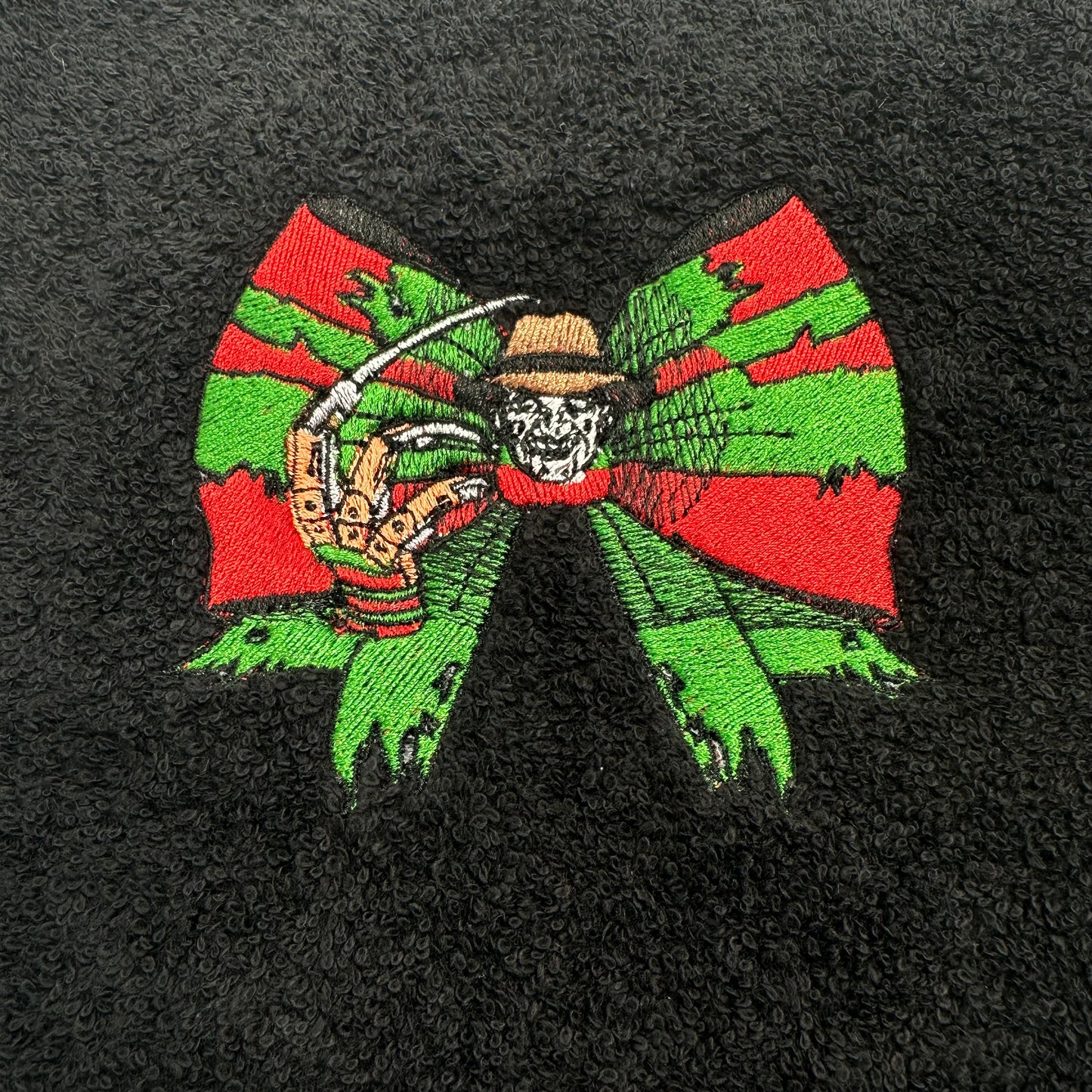 A close-up of a black textured fabric, likely a towel or blanket, featuring a colorful embroidered graphic of Freddy Krueger inside a large red and green bow.