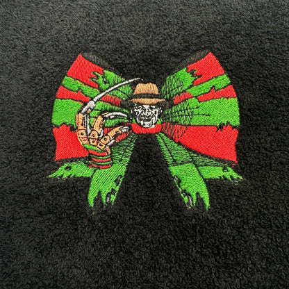 A close-up of a black textured fabric, likely a towel or blanket, featuring a colorful embroidered graphic of Freddy Krueger inside a large red and green bow.
