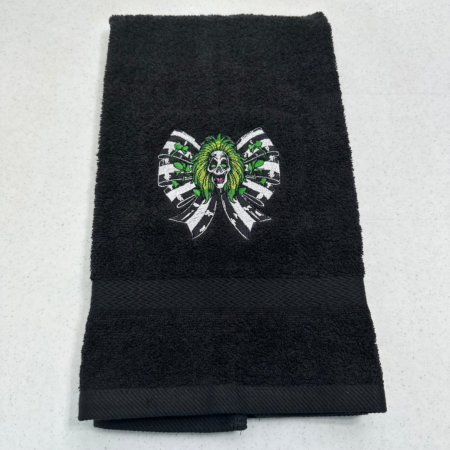 A black terry cloth hand towel with a central embroidered graphic of a green-haired beetlejuice surrounded by white and black ribbon-like shapes, laid flat on a light gray surface.
