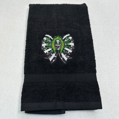 A black terry cloth hand towel with a central embroidered graphic of a green-haired beetlejuice surrounded by white and black ribbon-like shapes, laid flat on a light gray surface.