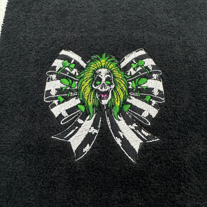 A close-up of a black textured fabric, likely a towel or blanket, featuring a central embroidered graphic of a beetlejuice with green hair, surrounded by a black and white striped bow with green shamrock accents.
