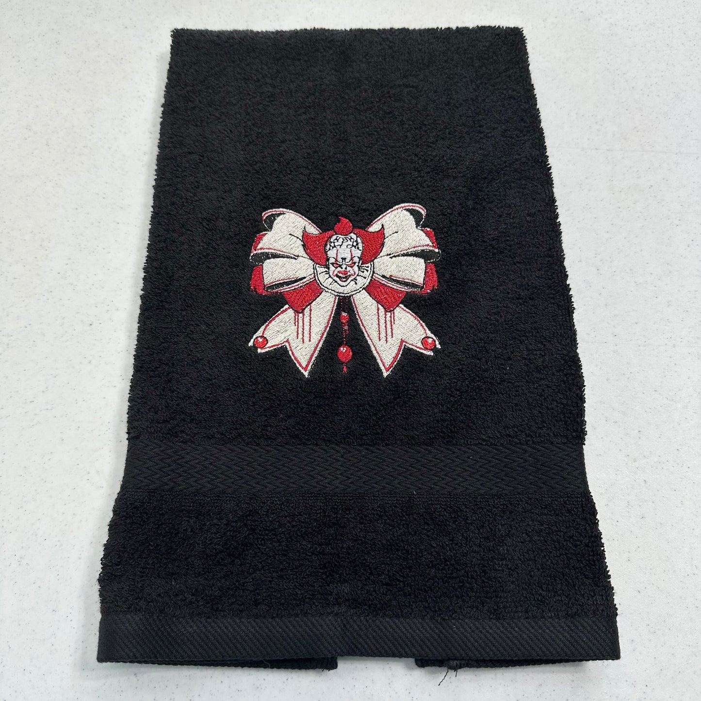A black terry cloth hand towel with a decorative embroidered bow with pennywise in red and white on the front, laid flat on a light gray background.