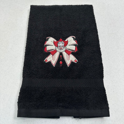 A black terry cloth hand towel with a decorative embroidered bow with pennywise in red and white on the front, laid flat on a light gray background.