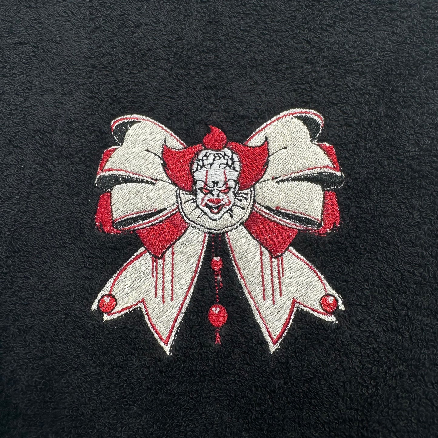 A close-up of a black textured fabric featuring a detailed embroidered design of Pennywise the Clown, styled as a large white bow with red accents and a creepy clown face in the center.