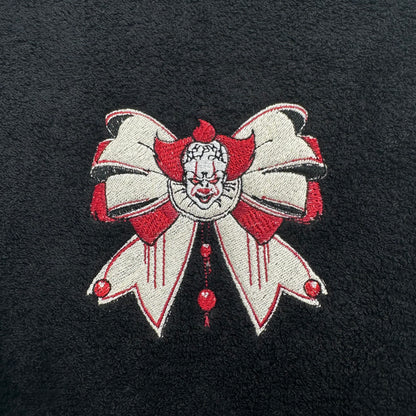 A close-up of a black textured fabric featuring a detailed embroidered design of Pennywise the Clown, styled as a large white bow with red accents and a creepy clown face in the center.