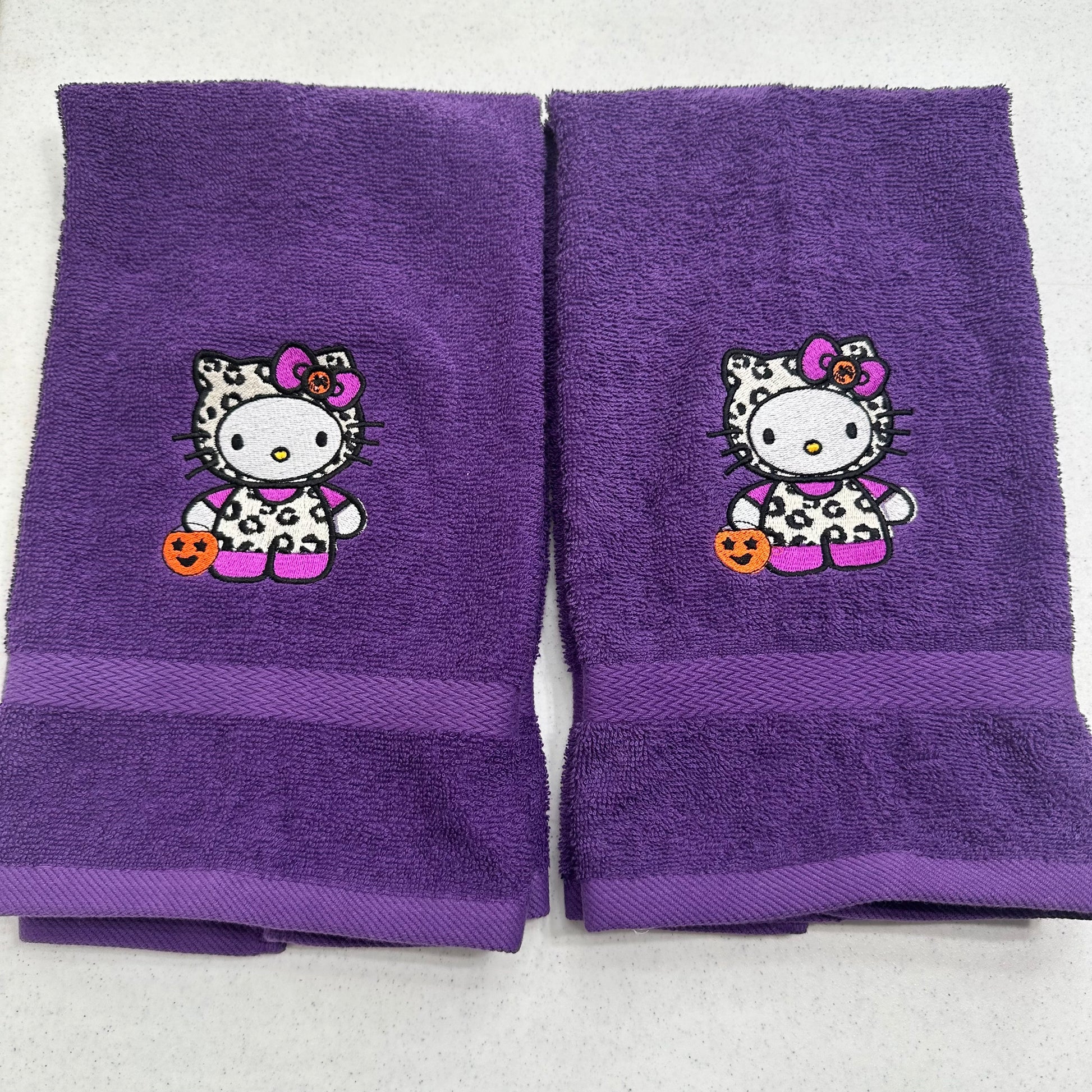 Two purple towels with embroidered Hello Kitty Halloween designs, displayed side-by-side on a light gray background.