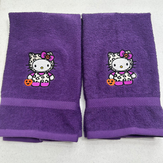 Two purple towels with embroidered Hello Kitty Halloween designs, displayed side-by-side on a light gray background.