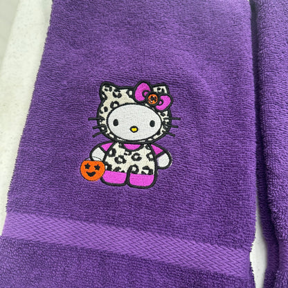 A purple terry cloth towel featuring an embroidered Hello Kitty character in a Halloween outfit with a leopard-print pattern, holding an orange pumpkin, and wearing a pink bow.