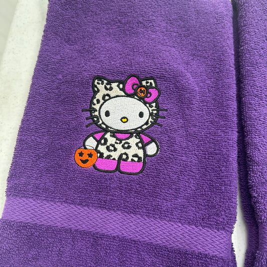 A purple terry cloth towel featuring an embroidered Hello Kitty character in a Halloween outfit with a leopard-print pattern, holding an orange pumpkin, and wearing a pink bow.