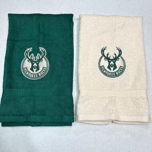 Two folded towels side by side on a light gray background. The left towel is dark green with a white embroidered Milwaukee Bucks deer logo, and the right towel is cream with a dark green embroidered Milwaukee Bucks deer logo.