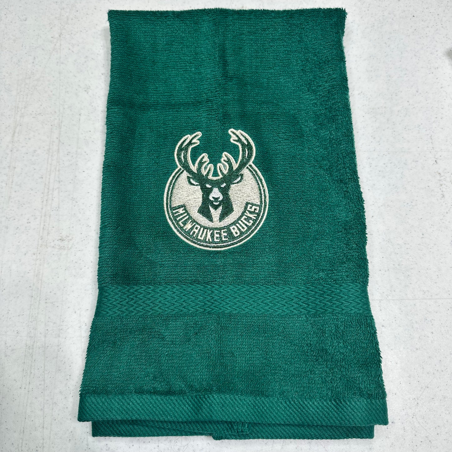 A dark green Milwaukee Bucks towel with the team logo embroidered in the center, laid flat on a light gray textured surface.