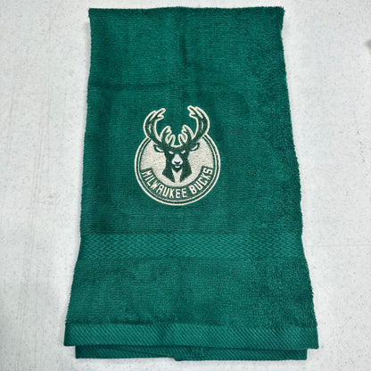 A dark green Milwaukee Bucks towel with the team logo embroidered in the center, laid flat on a light gray textured surface.