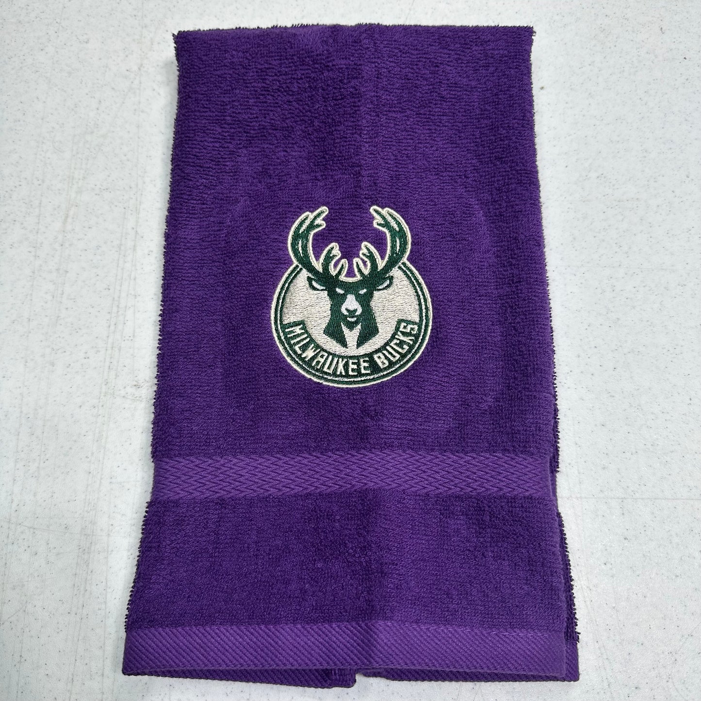 A purple Milwaukee Bucks towel with a green and white embroidered team logo in the center and a horizontal textured stripe near the bottom, laid flat on a light gray surface.