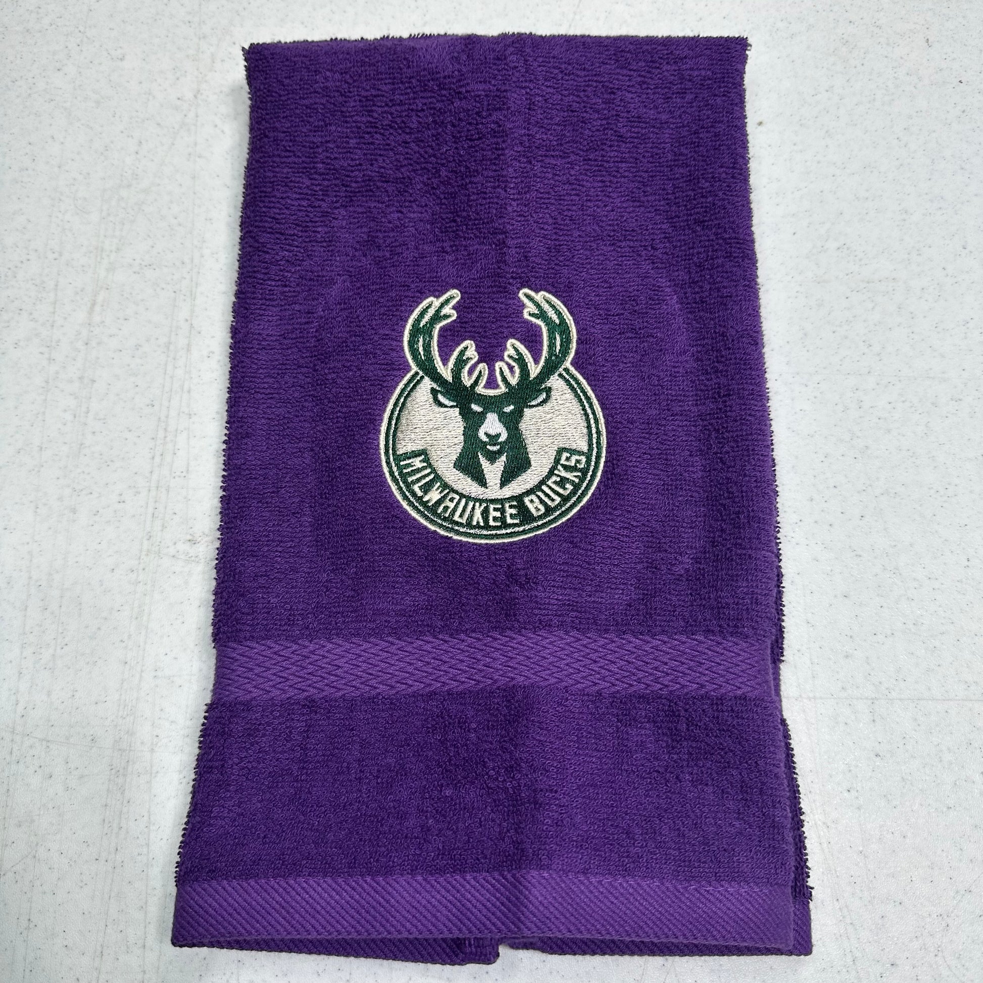 A purple Milwaukee Bucks towel with a green and white embroidered team logo in the center and a horizontal textured stripe near the bottom, laid flat on a light gray surface.