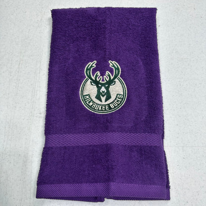 A purple Milwaukee Bucks towel with a green and white embroidered team logo in the center and a horizontal textured stripe near the bottom, laid flat on a light gray surface.