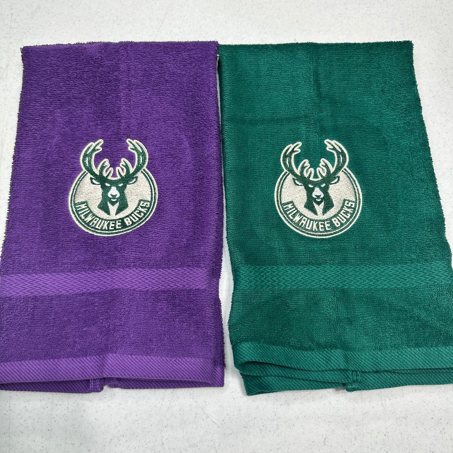 Two folded towels side by side on a light gray surface, one purple and one green, both featuring a white Milwaukee Bucks logo and a horizontal textured stripe near the bottom.